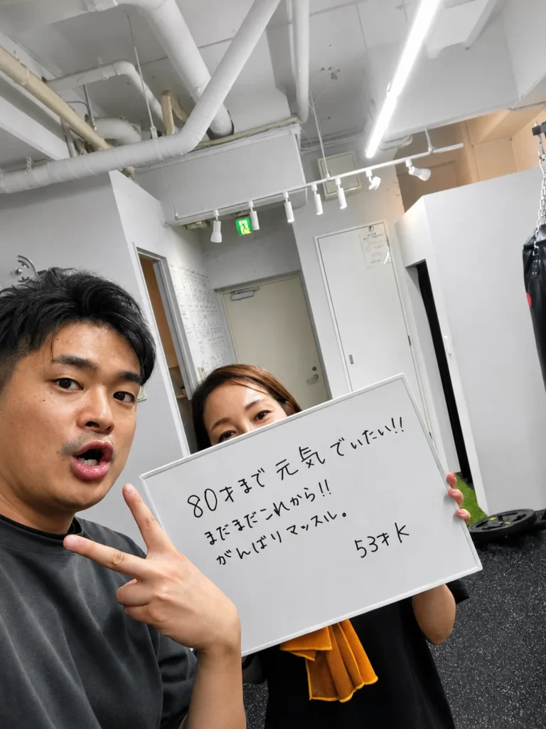 HaneGymでパーソナルトレーニングを続ける53歳の女性会員とトレーナーが、目標を書いたボードを持って記念撮影する様子