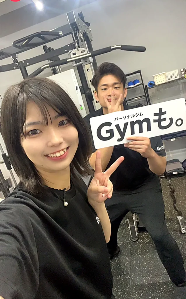 パーソナルジム「Gymも。」の店内で笑顔でピースする女性と看板を持つ男性スタッフ
