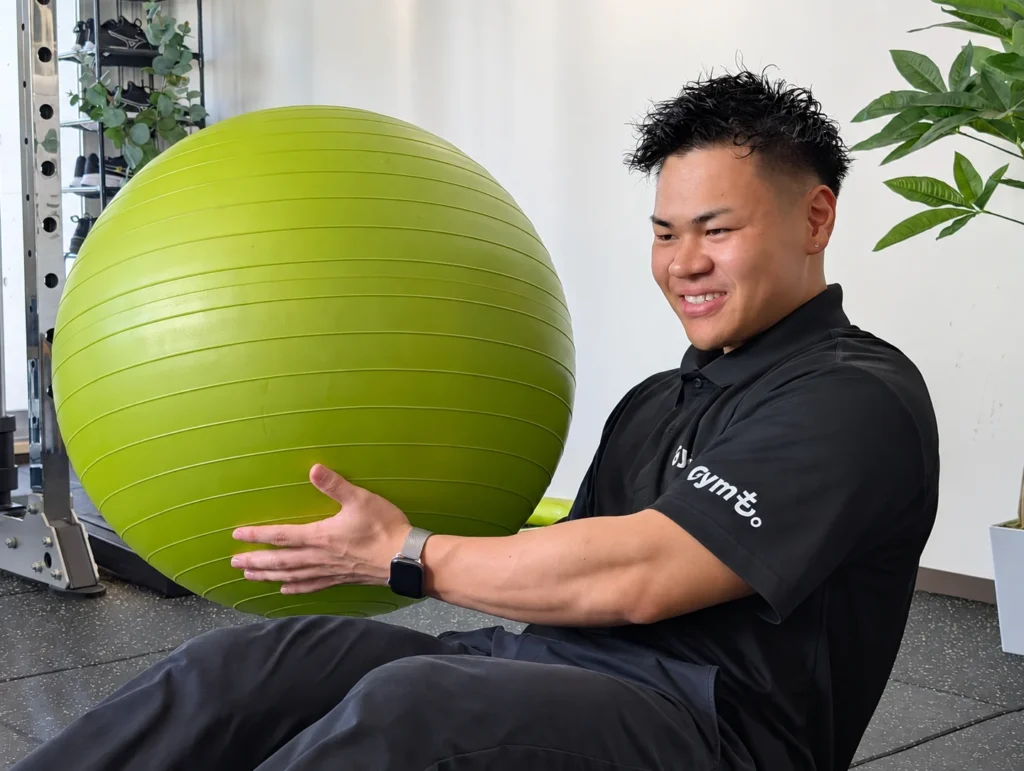 Gymも。の小川トレーナーが、座った姿勢で緑色のバランスボールを両手で持つ様子