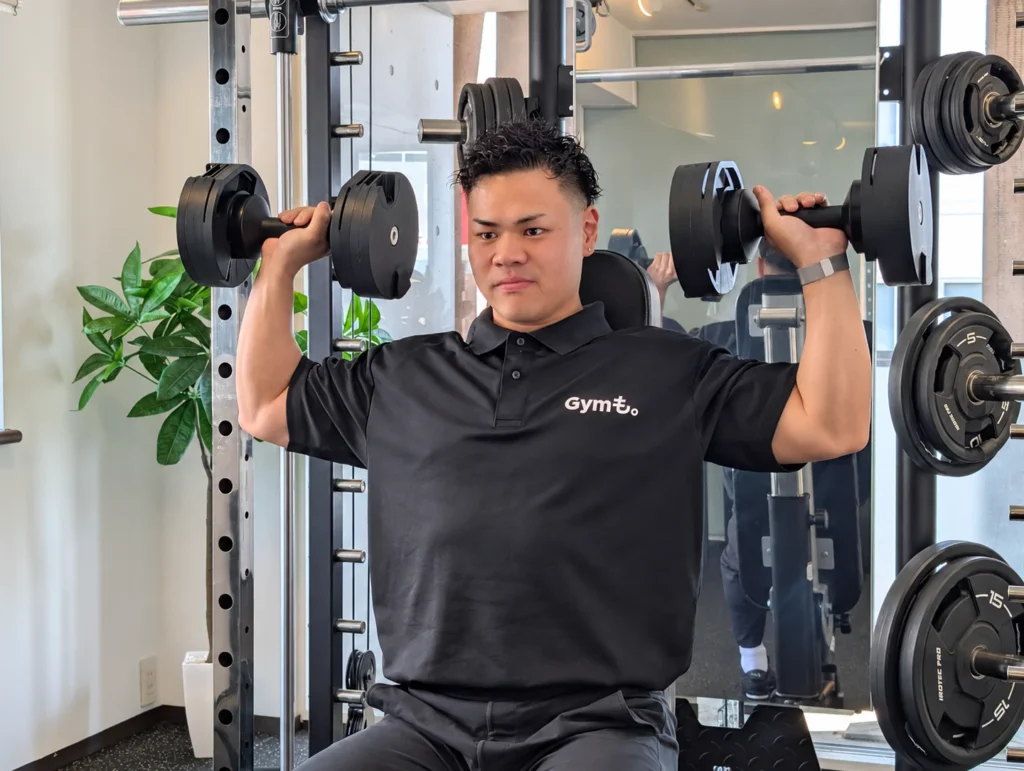 Gymも。の小川トレーナーが、両手にダンベルを持ってショルダープレスを行う様子