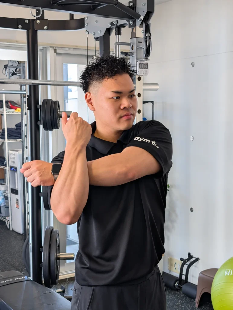 Gymも。の小川トレーナーが、片腕を胸の前に伸ばして肩のストレッチを行う様子