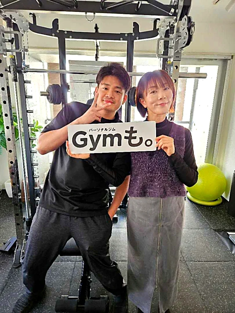 パーソナルジムGYMも。の店内でロゴボードを持つ男女2名の記念写真。トレーニング設備を背景に撮影。