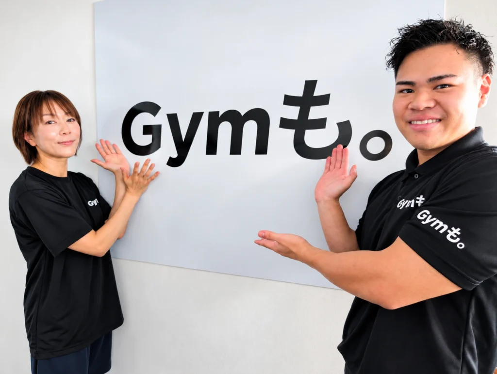 Gymも。志木店の小川トレーナーとお客様が、Gymも。のロゴの壁の前で笑顔を見せる様子
