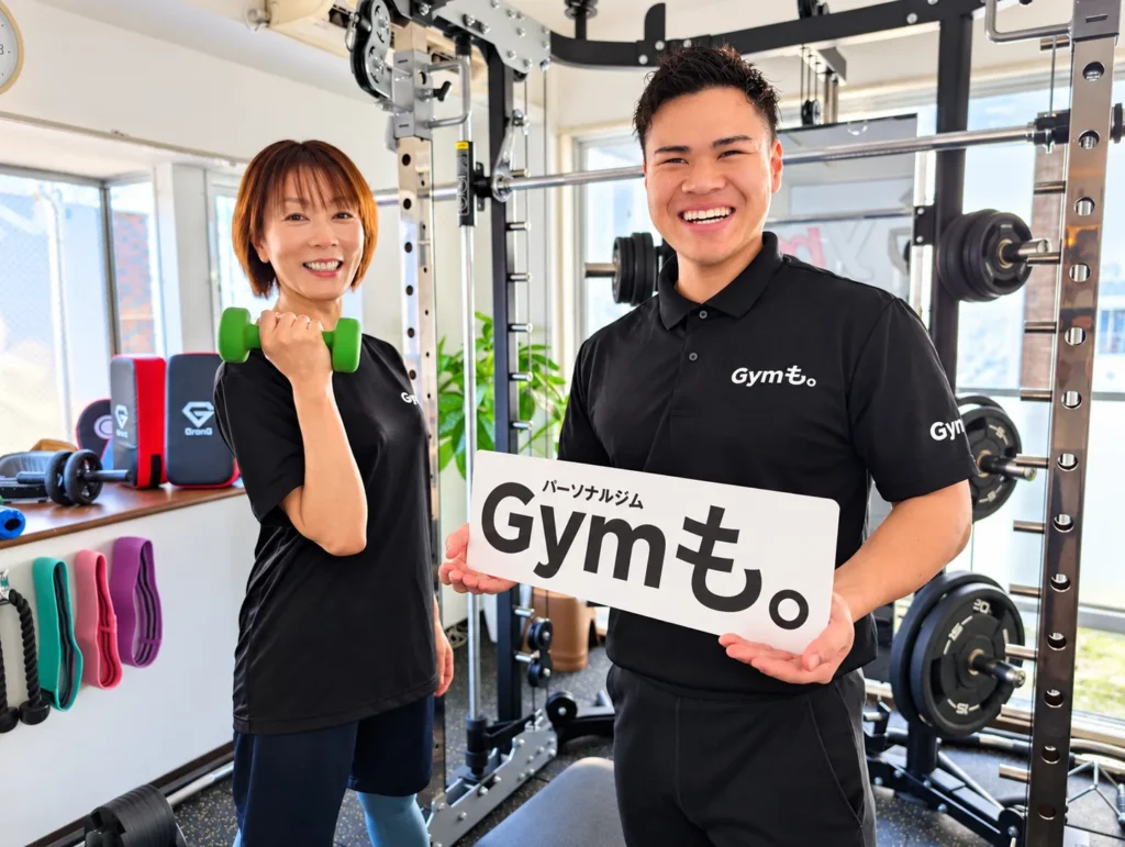 Gymも。志木店の小川トレーナーが、お客様と一緒にGymも。の看板とダンベルを持って笑顔を見せる様子