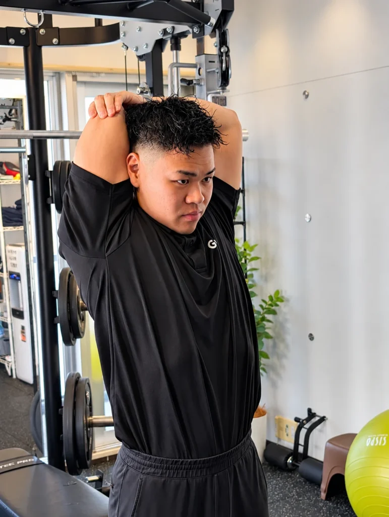 Gymも。の小川トレーナーが、片腕を頭の後ろに曲げて二の腕の裏側を伸ばす様子