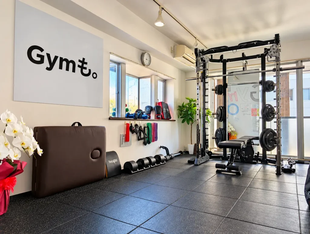 志木のパーソナルジムGymも。志木店の店内。Gymも。のロゴの壁とパワーラック、ストレッチ用のベッド